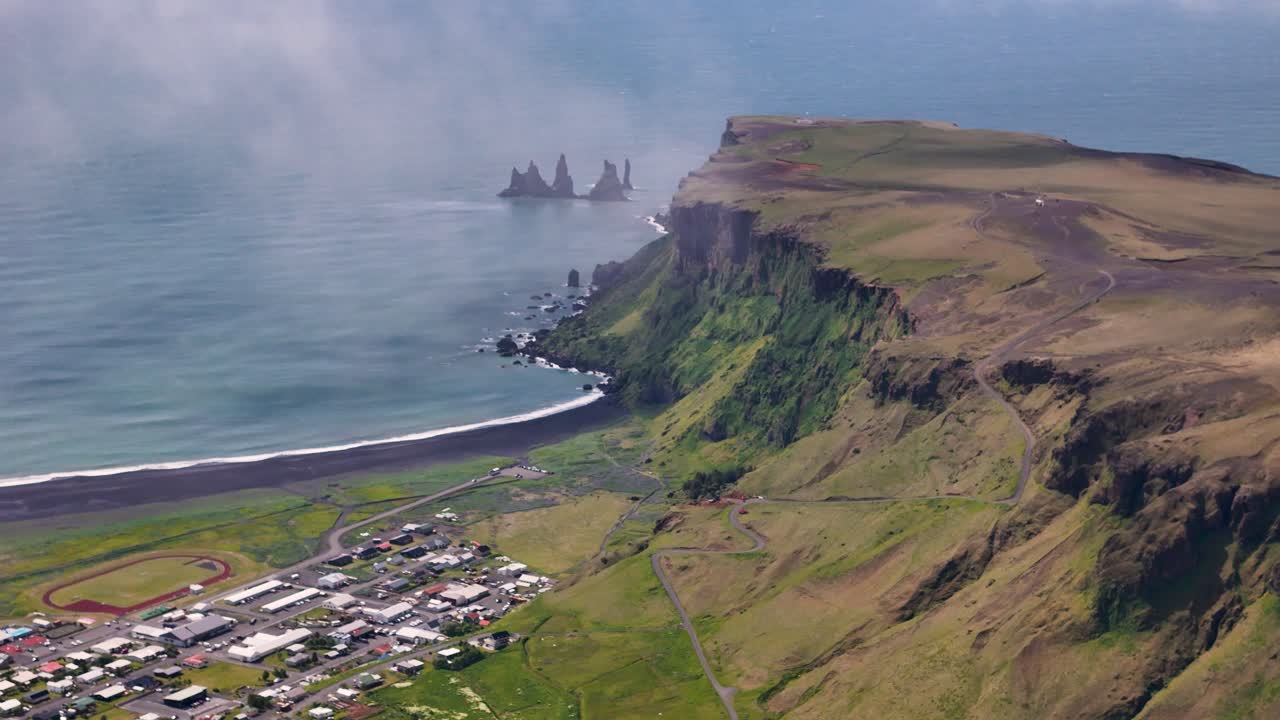 Drone revealing of green mountain side of Iceland during sunny day. Vik city at coast and black reynisdrangar rock formation in Iceland. Aerial wide shot.