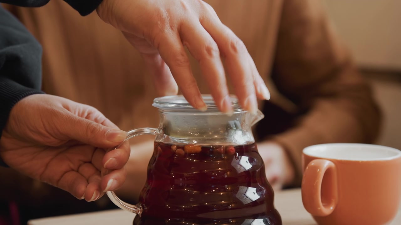 Primo piano di una persona che versa un liquido scuro da una caraffa di vetro in una tazza arancione con un'altra persona seduta sullo sfondo. La scena presenta un'illuminazione soffusa e toni caldi, concentrandosi sulle mani.