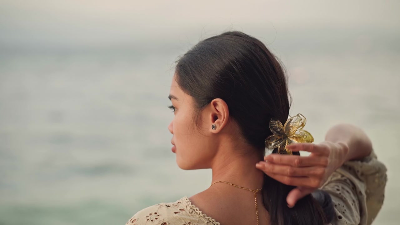 Donna con fiori guarda l'oceano. Serena donna asiatica osserva un paesaggio marino calmo sotto la luce del sole. Calma donna asiatica con accessorio floreale tra i capelli contempla una scena oceanica pacifica durante l'ora d'oro.