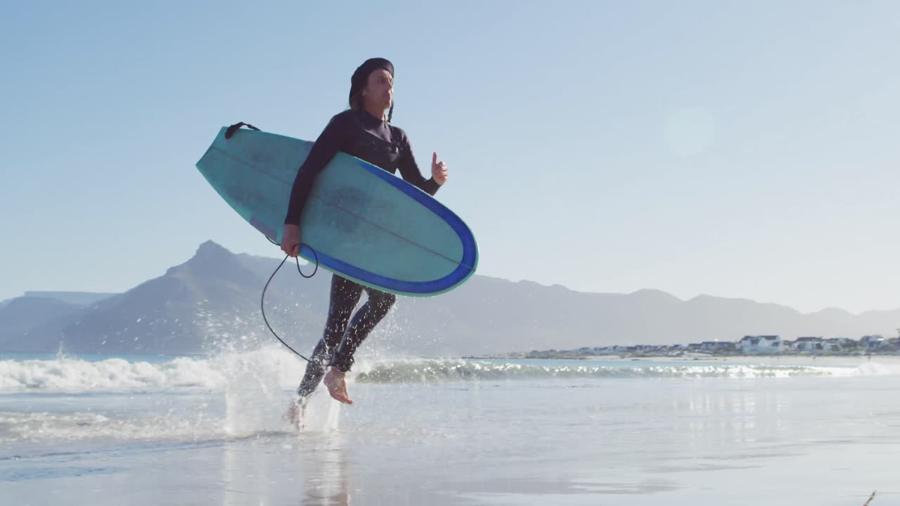바다에서  ⁇ 은 해변으로 달리는 서핑 보드를 들고 있는 웨이트스 ⁇ 을 입은 백인 남자의 비디오