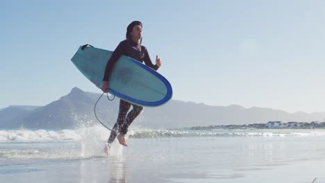 video de un hombre caucásico con rastas en traje de neopreno llevando una tabla de surf corriendo desde el mar a la playa soleada