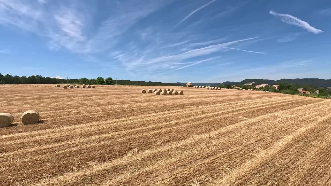 A smooth FPV drone video captures the serene landscape of a harvested cereal field dotted with neatly arranged hay bales. The scene highlights the expansive countryside under a partly cloudy sky.