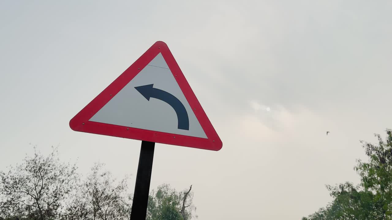 La cámara orbita alrededor de una señal de tráfico triangular que advierte de un giro a la izquierda en un poste, con borde rojo y flecha negra visible sobre un cielo pálido con árboles distantes en condiciones de luz diurna