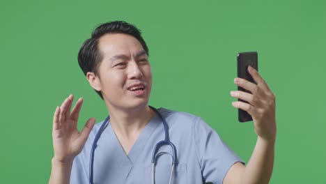 스테토스코프를 들고 있는 아시아 남성 의사가 손을 흔들고 병원에서 녹색 화면 배경에 서 있는 동안 스마트폰으로 비디오 통화를 하고 있다.