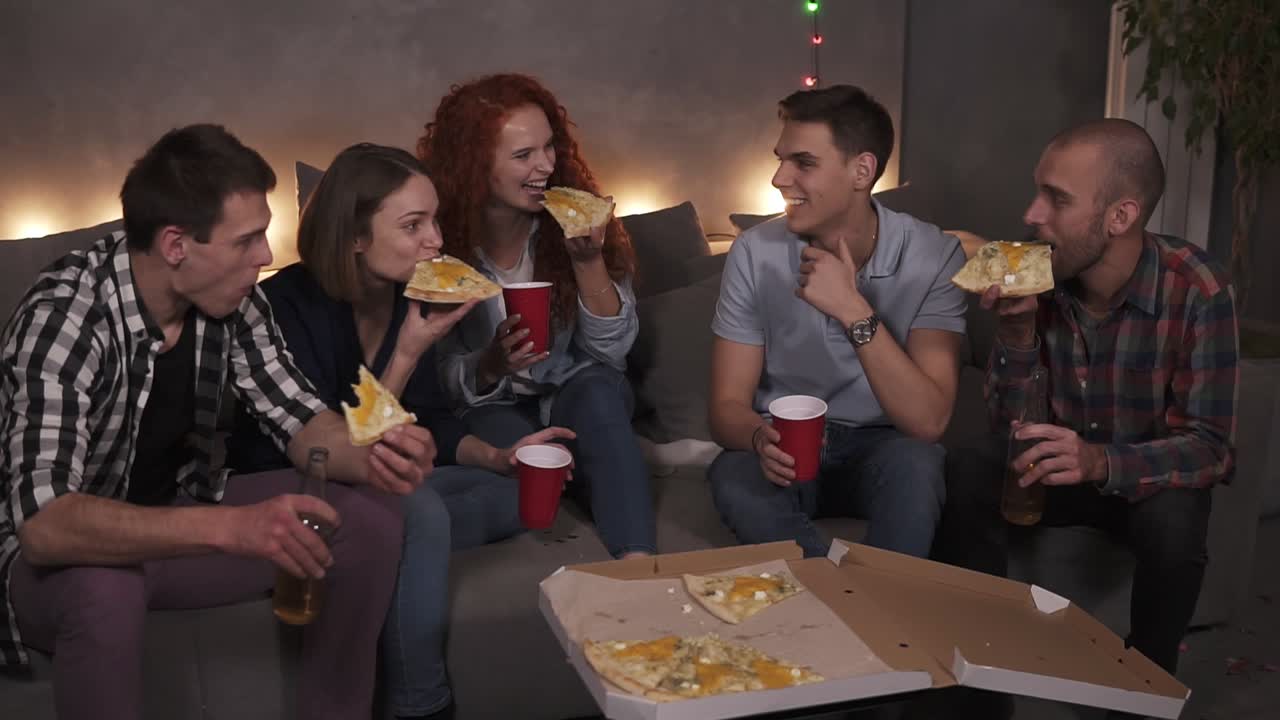 jóvenes hombres y mujeres alegres están comiendo pizza de queso, charlando relajándose durante una fiesta cubierta en un apartamento en una habitación de ático. una deliciosa pizza en caja de cartón es visible en la mesa, sosteniendo botellas y tazas con bebidas