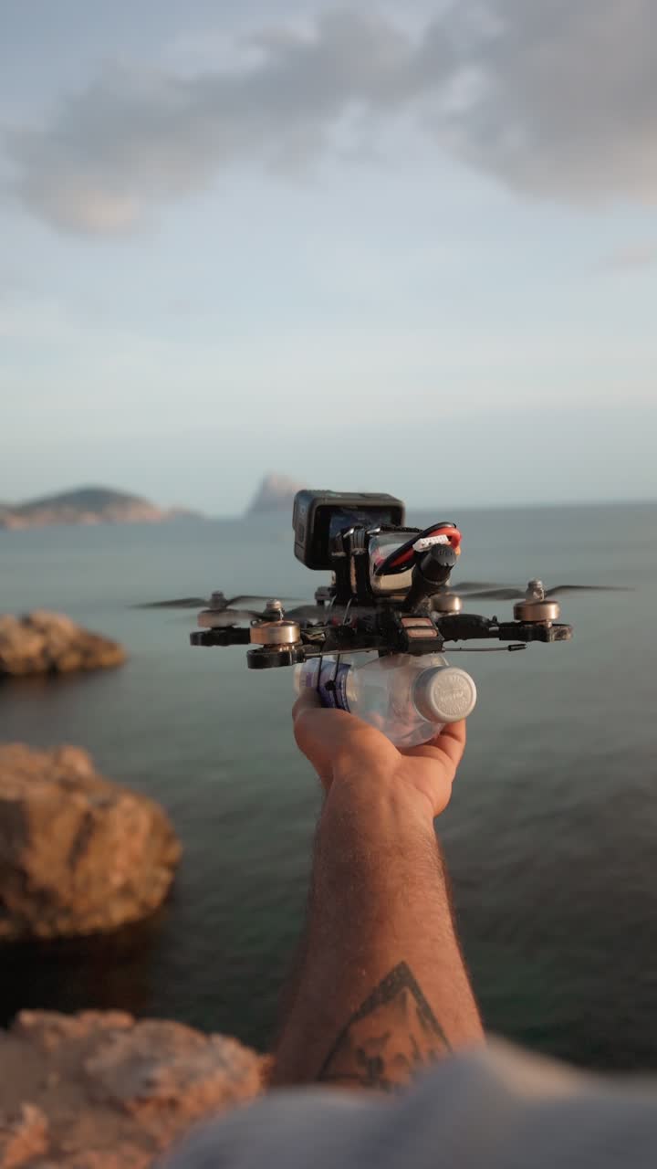 Close-up of a hand launching an FPV drone with a plastic bottle attached, by the sea with rocks in background