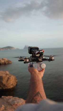 close-up of a hand launching an fpv drone with a plastic bottle attached, by the sea with rocks in background