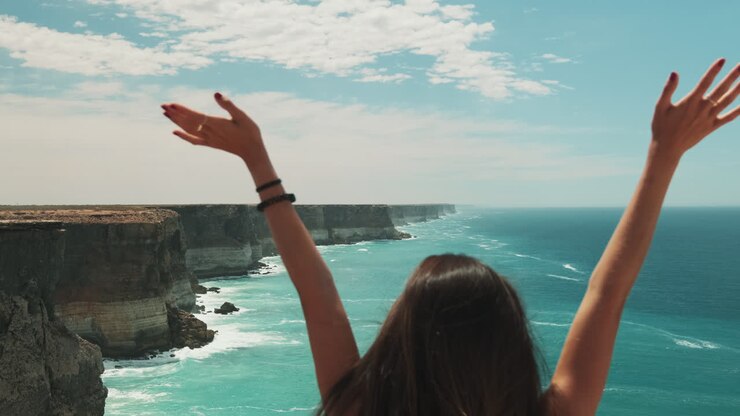 Mujer bonita levantando las manos con una épica vista del océano australiano de fondo, toma trasera.
