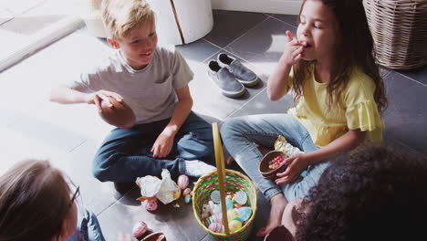 group of children sitting on floor at home eating chocolate eggs they have found on easter egg hunt