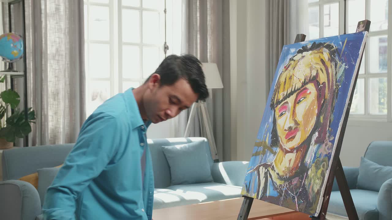 Medium Shot Of An Asian Artist Man Coming To Sit Down And Holding Paintbrush Mixed Colour Before Painting A Girl'S Hair On The Canvas
