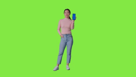 full length studio portrait of woman holding blue screen mobile phone towards camera against green screen