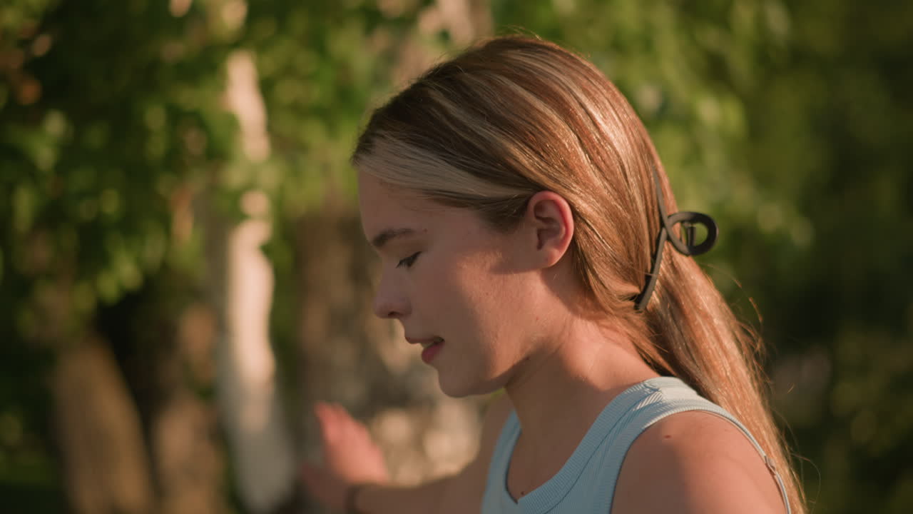 태양빛이 반사되면서 스케이트를 타는 것처럼 우아하게 움직이는 동안 미소 짓는 젊은 여자의 근접 측면 뷰, 머리카락은 나무의 자연적인 배경으로 뒤로 여 있습니다.