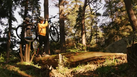 Una-Pareja-De-Ciclistas-De-Montaña-Llevando-Una-Bicicleta-Mientras-Caminaba-Sobre-Un-Tronco.