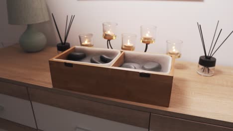 close-up of black massage stones in a wooden box, set against a backdrop of beautiful burning candles on a dresser