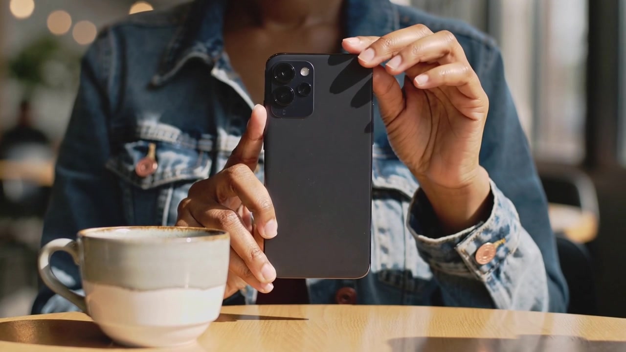 Reaching into jacket, woman in denim pulling black phone out at cafe, inspecting camera near mug