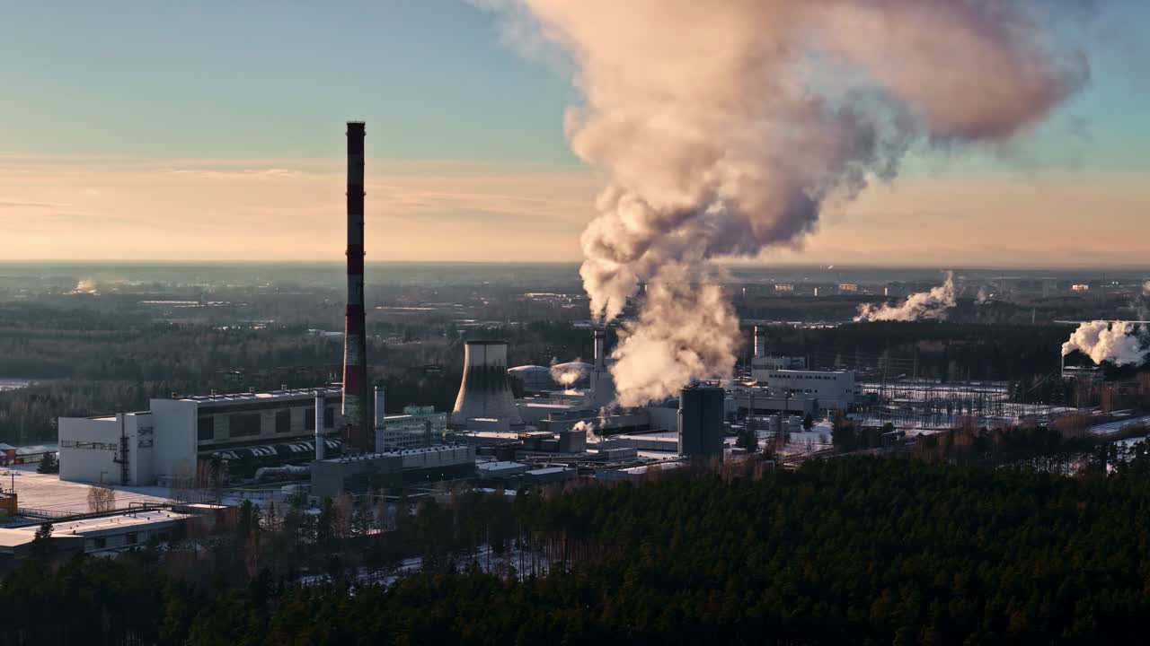 Centrale elettrica industriale con alte ciminiere che rilasciano pennacchi di vapore e fumo nel cielo, circondata da un paesaggio rurale e una foresta