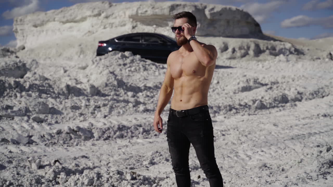 Athlete with muscular body in white mountains. Handsome sportsman without shirt in sunglasses stands on canyon on black car background.