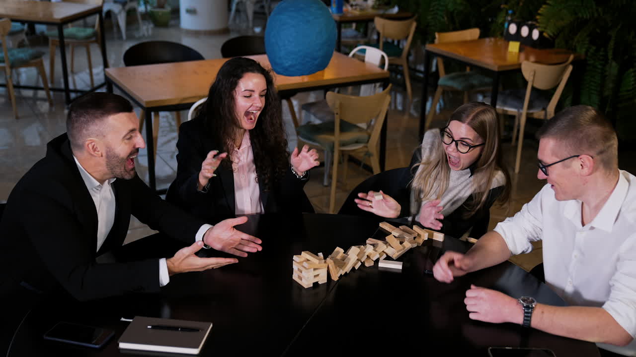 Adult friendly people pay jenga game building a tower from wooden blocks. Blonde girl in glasses pulls a brick and breaks a pile. View from top.