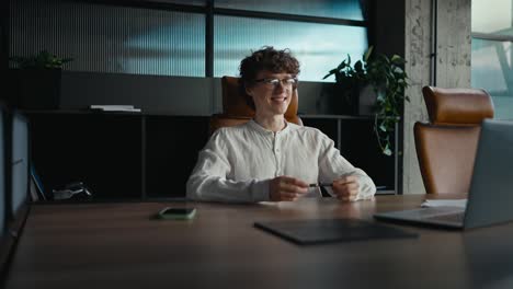 Side-view-of-a-happy-young-guy-with-curly-hair-wearing-glasses-in-a-white-shirt-communicates-via-video-conference-using-a-gray-laptop-while-sitting-at-a-table-in-the-office