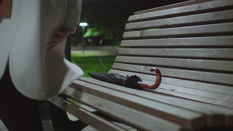 back view of lady dropping umbrella on wooden bench then sitting calmly as she opens book to read under night park light, surrounded by greenery with soft streetlight bokeh in background