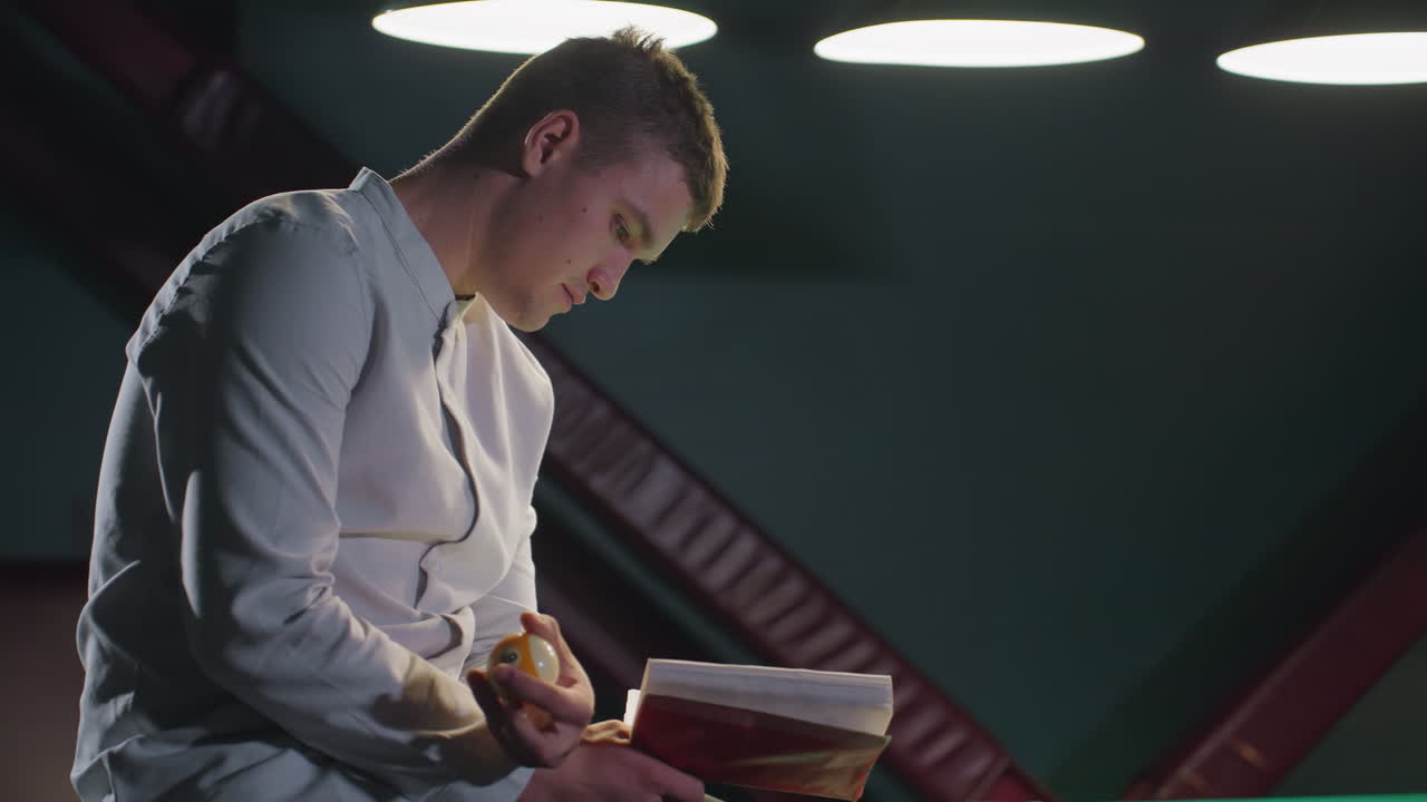 primer plano de un joven con camisa blanca sentado en una mesa de billar verde, leyendo un libro mientras tira una pelota de billar en la mano. la expresión enfocada bajo una iluminación tenue crea una atmósfera reflexiva y relajada
