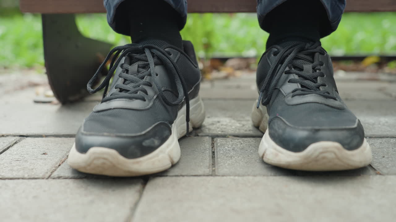 Nærbillede af sneakers på fortov under en bænk, mand der sidder stille med samlede fødder, slidte sorte kondisko, åbne snørebånd, tekstureret sål, parkgræs i baggrunden, dæmpet dagslys, eftertænksom stemning, ventende, urbant