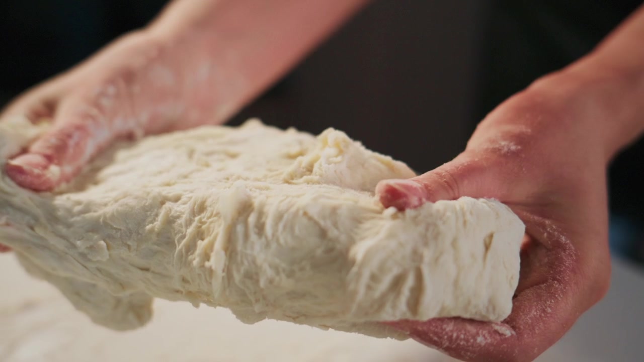 Mani impastano pasta morbida su una superficie infarinata. Le dita premono l'impasto, mostrando consistenza ed elasticità. La persona è concentrata, il tocco delicato suggerisce cura e pazienza. Ideale per cucina, pasticceria