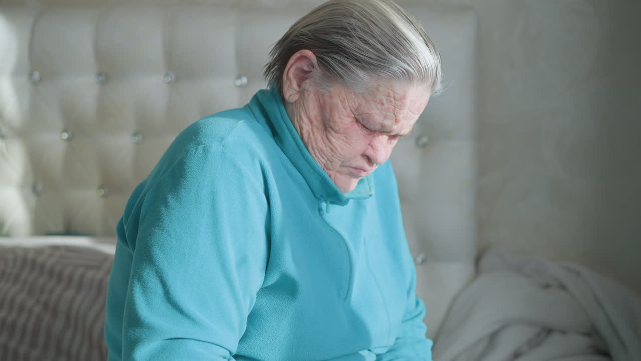 Old woman in green feeling discomfort in back, seated on bed, massaging lower back, looking tired. Senior care, wellness, physical health, elderly people dealing with back pain, aging process