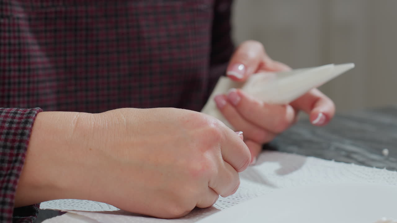Close-up of baker holding white piping bag in one hand while using toothpick in other hand to adjust icing on cookie, focusing on careful decoration with precision on tissue paper surface