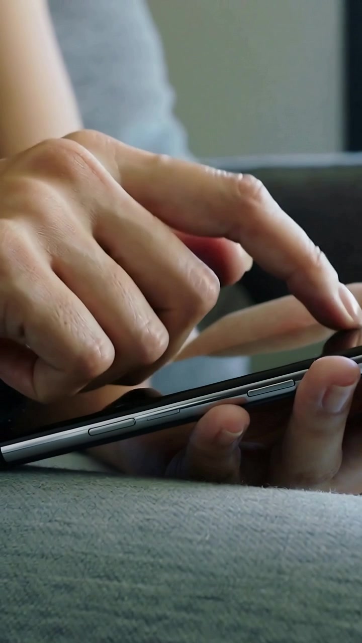 Vertikální video: Klepající ukazováček se dotýká obrazovky telefonu, ruce přejíždějí po gauči v šedém tílku.