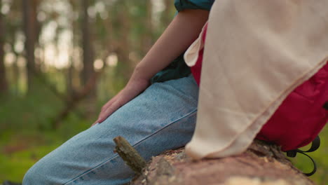 side view of hiker resting on fallen tree with red bag and cloth draped over it in peaceful forest setting,dressed casually in jeans, hand resting on leg