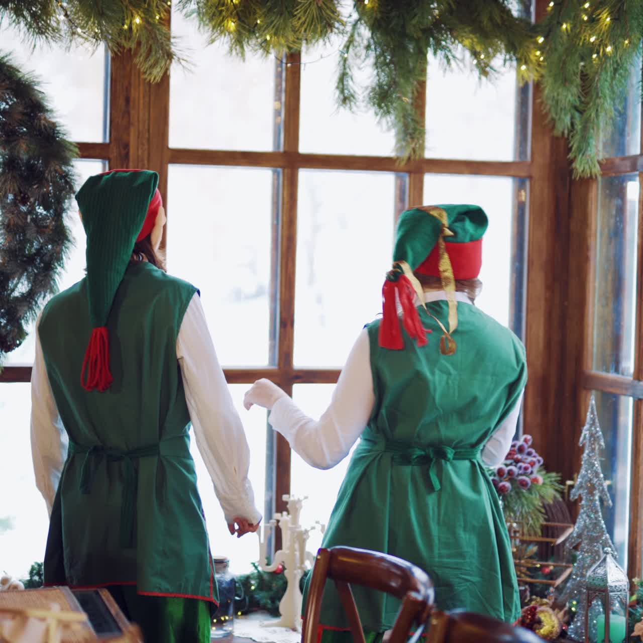 Joyful elves in green costumes are viewing the landscape from the window on the background of New Year decorations and Christmas wreath with a toy at day of New Year