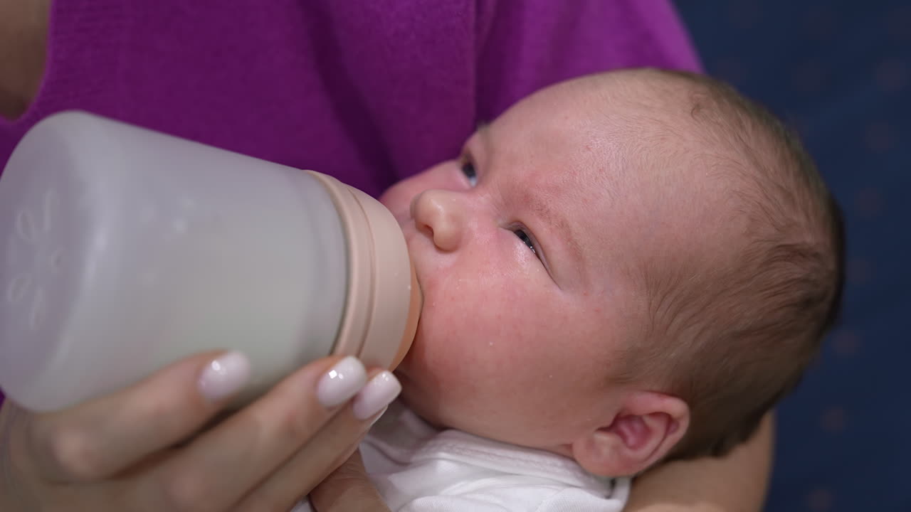 Newborn baby fed from a bottle lying in mother's hands. Baby boy screwing up his eyes against the light while being fed. Close up.