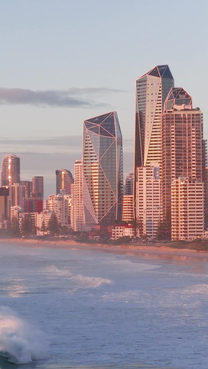 A vertical drone shot capturing modern city towers and rolling waves during golden hour.