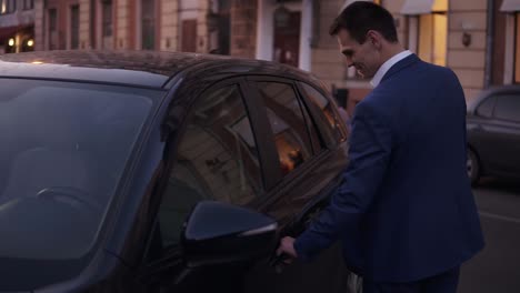 slow motion young man walk sitting in his new black car on street. smiling businessman in elegant blue suit opens the door of