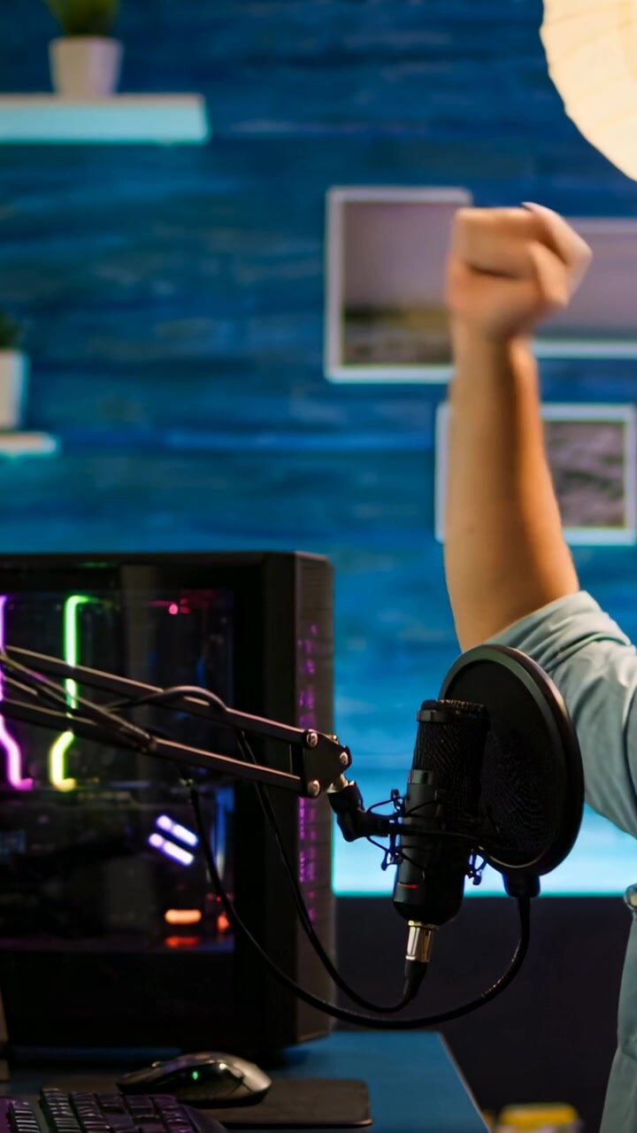 Video verticale: Un gamer con una maglietta blu alza il pugno destro ed esulta al microfono sul set, con un PC RGB e spazio per il testo.