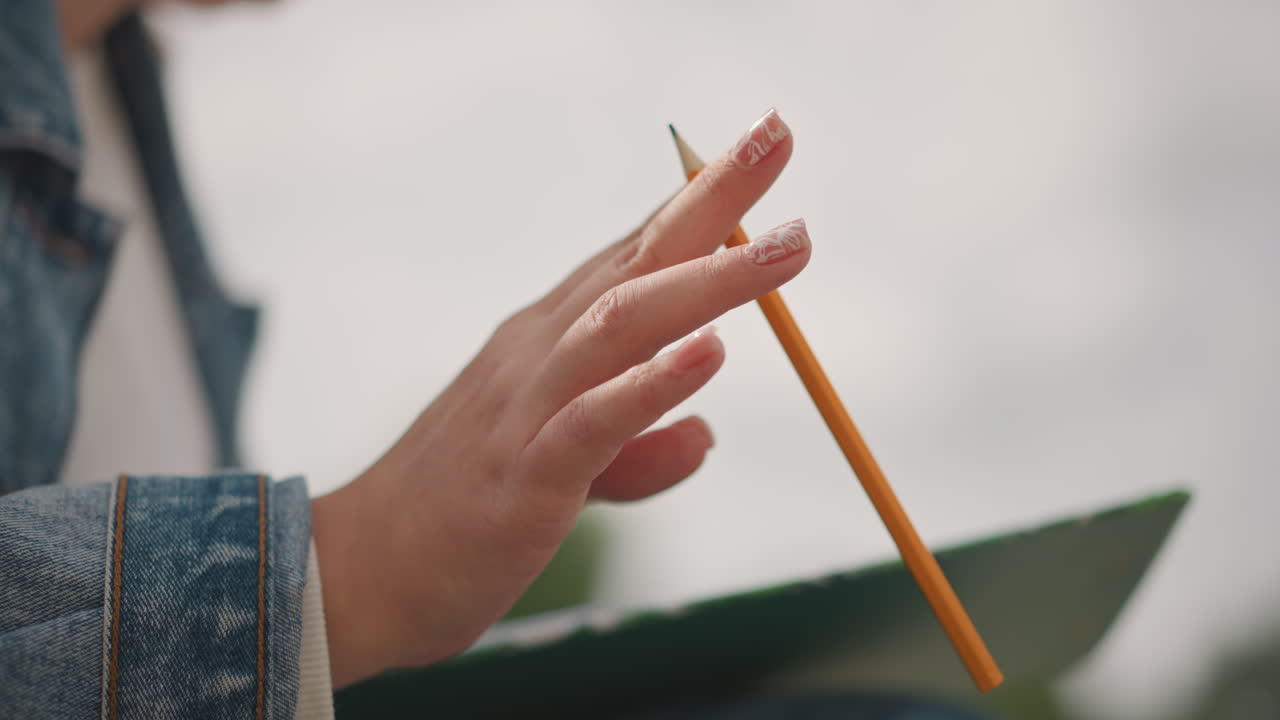 Pencil poised over clipboard, denim sleeve, delicate nails student sketcher pausing to observe composition, overcast light, soft background greenery, slow thoughtful gesture captured in shallow focus