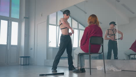 boy removes belt wraps strap around neck then tosses it aside before seated woman wearing cap glasses harness then pulls down trousers in mirrored dance studio with baton and discarded cloth on floor