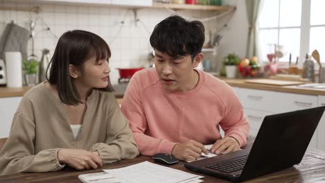 unhappy chinese married couple calculating budget on calculator in dining room at home. the surprised man holding his head worrying about lack of money to pay the loan