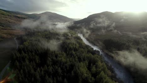 aerial drone footage slowly descending over a dark forest of conifer trees towards a river while shafts of light illuminate low hanging cloud in the treetops at sunset