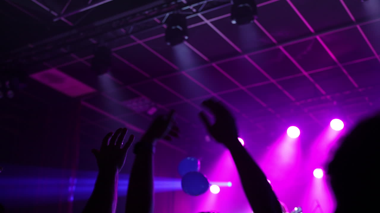 Enthusiastic music fans raising hands, creating energetic crowd atmosphere with colorful stage lighting illuminating silhouetted bodies during vibrant live performance