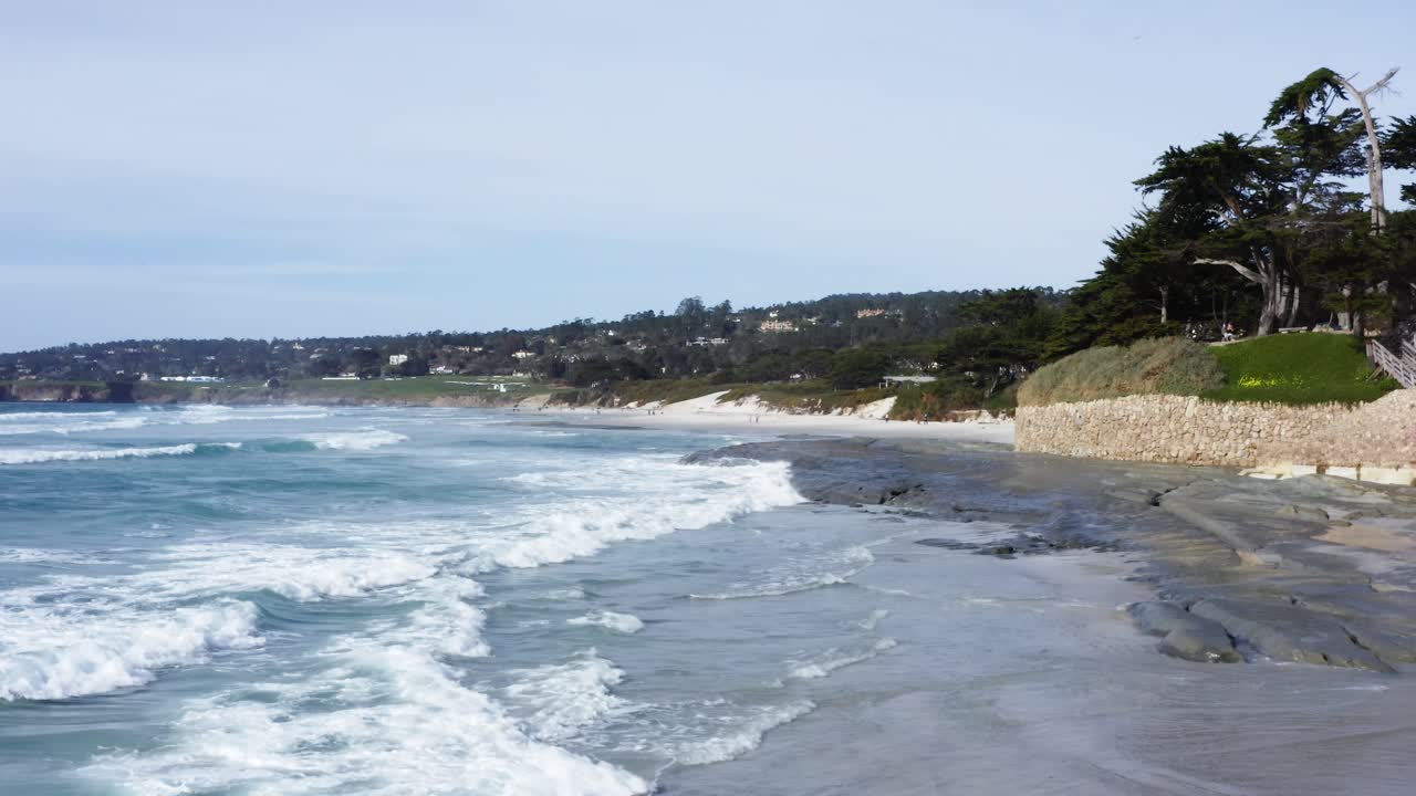 Aerial Flyover of Ocean waves rolling over exposed rocks at Carmel-by-the-Sea Beach with Pebble Beach Homes and Golf Course in background