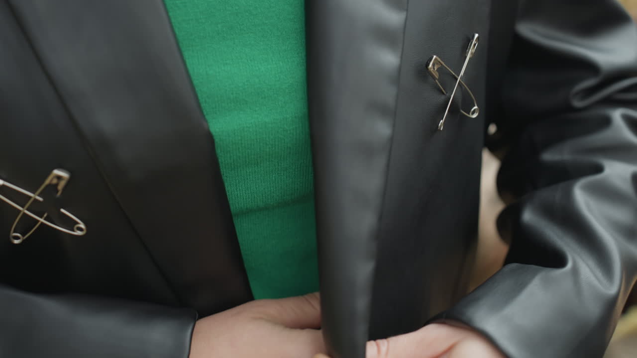 donna che tiene in mano un grande coltello seghettato all'aperto vicino a una foresta, giacca di pelle con chiusure in metallo e maglione verde visibile, primo piano sulle mani che stringono la lama, foglie autunnali e catasta di legna sullo sfondo per creare