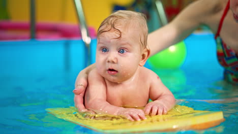 beautiful blue-eyed caucasian baby lies on the board in the swimming pool. woman is moving the kid by the pool.