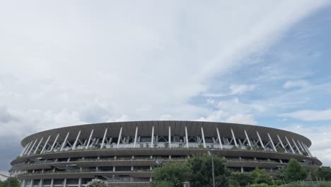 a wide, low-angle shot of the national stadium, featuring its modern architecture and green trees under a cloudy sky