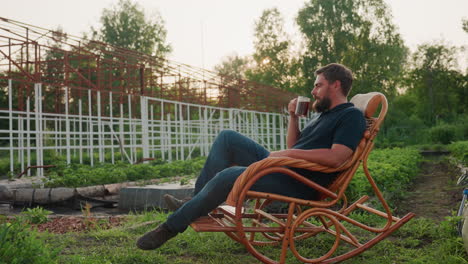 man seated in wooden rocking chair in garden greenhouse holds steaming herb tea cup poised for sip under soft evening light with lush plants and tranquil atmosphere gentle sunset breeze
