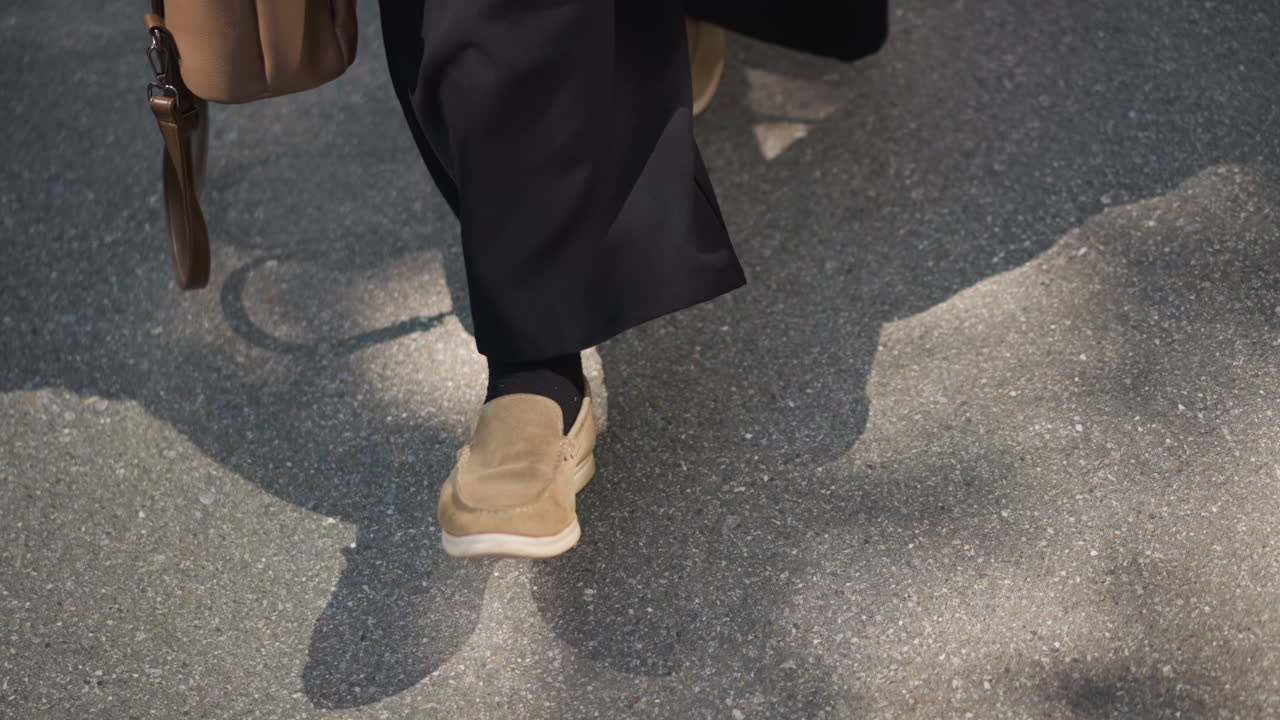Front leg view of girl walking outdoors holding brown handbag, soft sunlight casting shadow on pavement, gentle movement and casual shoes creating calm atmosphere of everyday