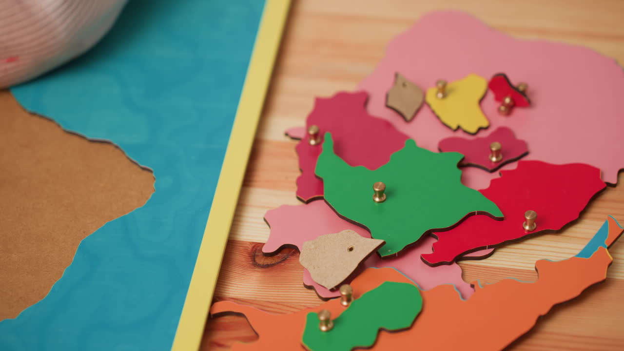 Primer plano de la mano de un bebé que sostiene una pieza de un puzle con la forma de un continente de un tablero de mapas Montessori; piezas de geografía de madera de colores apiladas sobre la mesa; aprendizaje de la motricidad fina, coordinación, práctica de colocación