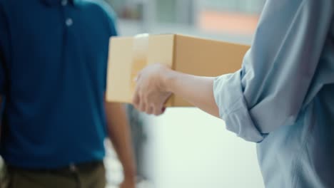 close up parcel in hand of blue uniform delivery man to customer front of the house with good service from shopping online. courier man delivering a cardboard box postal package to destination.