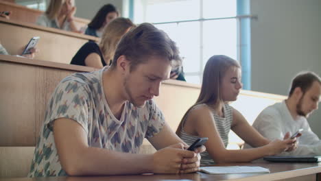 Eine-Gruppe-Von-Männern-Und-Frauen-In-Einem-Großen-Universitätspublikum-Schaut-Schweigend-Auf-Die-Bildschirme-Ihrer-Smartphones-Und-Schreibt-Chatnachrichten.-Jeder-Einzelne-Schaut-Auf-Den-Bildschirm-Seines-Geräts,-Ohne-Auf-Das-Geschehen-In-Der-Realen-Welt-Zu-Achten.